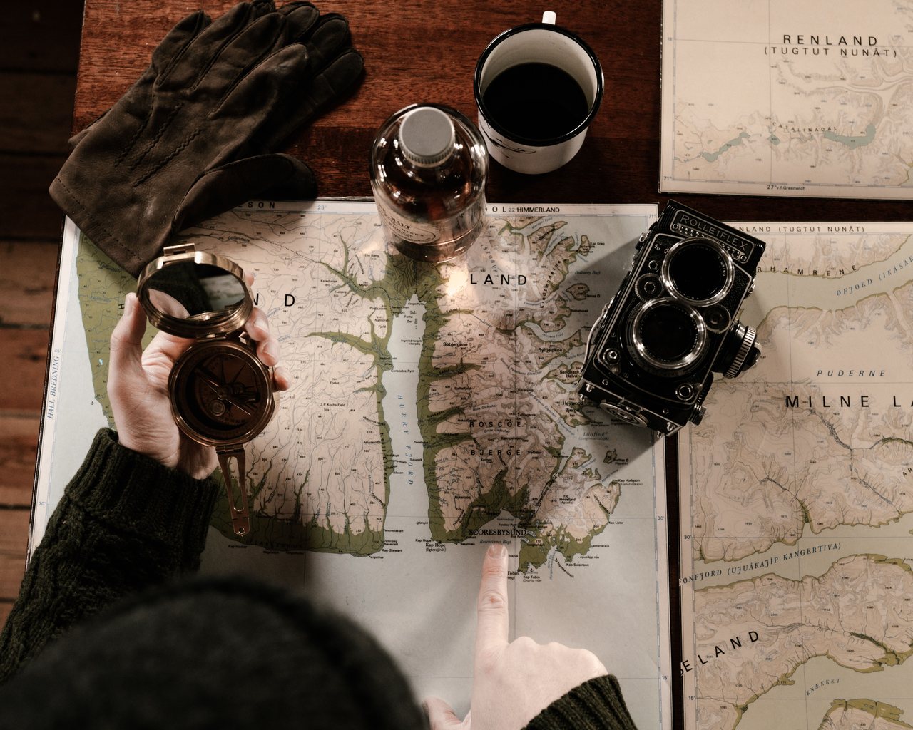 An overhead view of a map spread on a wooden table, with a brass compass in one hand, a finger marking a spot on the map, a vintage camera, leather gloves and a mug of coffee.